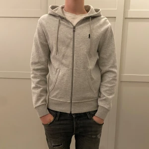 Grå hoodie från Polo Ralph Lauren - Snygg grå hoodie från Polo Ralph Lauren i storlek XS. Tröjan har huva med snörning, dragkedja framtill och två fickor. Liten broderad logga på bröstet. Mjuk bomullsmix som är skön att ha på sig. Perfekt för chill dagar. Nypris är ca 2000 kr! Passar upptill 180 cm 