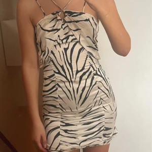 Zebramönstrad kort klänning med smala band - Snygg kort klänning med zebramönster i svart och beige. Klänningen har smala axelband, omlott-detalj framtill och en öppen rygg