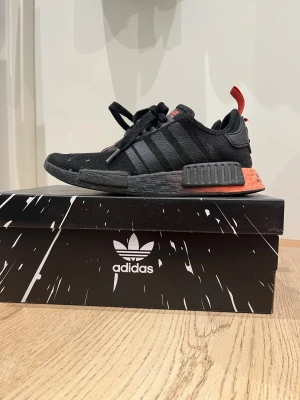 Svarta Adidas NMD sneakers - Snygga svarta Adidas NMD sneakers med klassiska tre ränder på sidan och en cool röd detalj på hälen. Skorna har en platt sula och snörning framtill. Materialet är syntet och textil, vilket gör dem både lätta och bekväma. Perfekta för dig som gillar streetstyle.