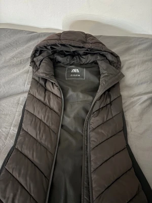 Svart väst från Zara - Svart väst från Zara med huva och quiltad design. Västen har dragkedja framtill och är tillverkad i syntetmaterial. Perfekt för lager-på-lager och har en stilren look med diskreta diagonala sömmar. Passar dig som gillar streetwear och vill hålla värmen.