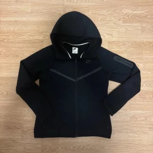 Nike Tech Fleece  - Svart Nike Tech Fleece från Nike i storlek XS. Den har hel dragkedja, huva och diskret Nike-logga på bröstet. Snygga detaljer med svarta linjer över bröstet och en ficka med dragkedja på ena ärmen. Perfekt för chill eller träning.