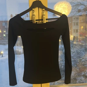 Svart långärmad topp med cut-out - Snygg svart långärmad topp med en cool cut-out detalj vid halsen. Toppens tighta passform och stilrena design gör den perfekt för dig som gillar minimalistiska plagg med en twist. Materialet känns mjukt och stretchigt mot kroppen.