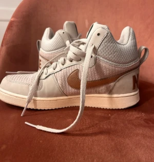 Nike sneakers med kopparfärgad swoosh - Snygga Nike sneakers i high-top modell med ljusgrå och beige mesh och skinnpaneler. Kopparfärgad swoosh och NIKE-text baktill ger en unik touch. Skorna har snörning och platt sula, perfekt för dig som gillar streetstyle och vill sticka ut.