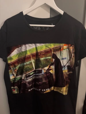 Svart Bastille t-shirt - Cool svart t-shirt från Bastille med stort färgstarkt tryck framtill som visar en bil och en Batman vid en bensinstation. Rund hals och korta ärmar. Tillverkad i mjuk bomull, made in Italy. Perfekt för dig som gillar unika motiv och streetstyle.