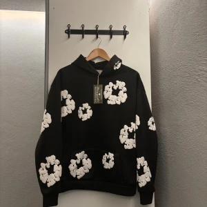 Denim Tears hoodie svart - Den är ganska oversized så passar för folk som tar på sig M också. Svart hoodie från Denim Tears med stora vita blommor tryckta över hela tröjan. Klassisk huva, känguruficka och relaxed fit. Perfekt statement-plagg för dig som vill sticka ut med en unik och trendig stil.