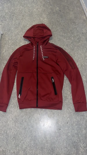 Vinröd hoodiejacka från McKenzie - Snygg vinröd hoodiejacka från McKenzie med huva och svarta dragkedjor på fickorna. Jackan har vita snören i huvan och logga på bröstet samt diskret tryck på ärmen. Perfekt för dig som gillar sportig och avslappnad stil.