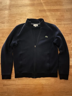 Lacoste zip tröja - Snygg marinblå ziptröja från Lacoste Sport med klassisk krokodillogga på bröstet. Tröjan har hög krage, dragkedja hela vägen och två sidofickor. Materialet är mjukt och skönt, perfekt för chill eller träning. Passformen är normal och ärmarna är långa.
