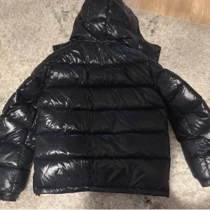 Svart pufferjacka från Moncler - Säljer en svart, glansig pufferjacka från Moncler med huva och dragkedja framtill. Jackan har två sidofickor med dragkedja och en ficka med Moncler-logga på ärmen. Perfekt för kalla dagar med sin tjocka vaddering och snygga look.