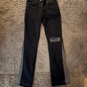 Flared svarta jeans med slitning - Flared jeans från MAN i svart denim med gråa paneler längs utsidan av benen. Framsidan har en snygg sliten detalj vid knät och råa kanter vid bensluten. Klassisk femficksmodell och knappgylf. Perfekt för dig som gillar en unik look. 29x30(31) ish