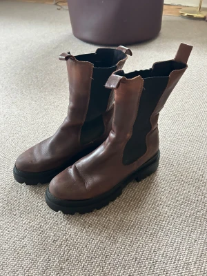 Tamaris Bruna chunky chelsea boots i skinn - stovlar - Säljer ett par bruna Tamaris boots med grov, svart platt sula och rund tå. Bootsens ovandel är i skinn med elastiska svarta paneler på sidorna och dragflikar upptill. Perfekt för dig som gillar robust stil och vill ha något som sticker ut. De är använda men i gott skick, utan skador, och har nyligen blivit rengjorda och behandlade med lädervård för att fräscha upp materialet. Innan leverans kommer de även att få ett extra lager vattenavvisande skydd, så de är redo för kalla och fuktiga dagar.