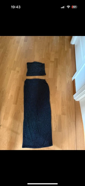 Glittrig marinblå tvådelad långklänning - Säljer en snygg tvådelad långklänning i marinblått med glittrigt tyg. Toppen är axelbandslös och kjolen är lång och rak. Perfekt för dig som vill sticka ut med en cool och trendig look. Materialet är mjukt och stretchigt syntet.