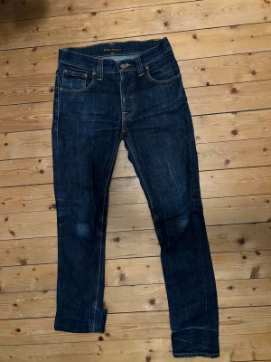 Nudie Jeans - Snygga mörkblå jeans från Nudie Jeans med klassisk femficksdesign och kontrastsömmar i orange. Modellen har rak passform och normal midja, med läderpatch och logga bak. Tillverkade i slitstarkt bomullsjeansmaterial, perfekta för en cool och avslappnad stil. 