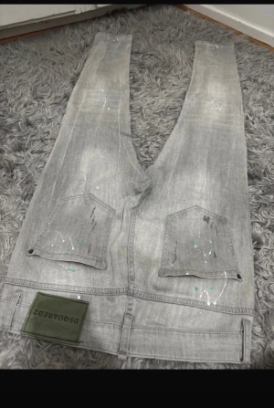Grå jeans från Dsquared2 med färgstänk - Säljer ett par grå jeans från Dsquared2 med coola färgstänk och slitningar på både fram- och baksida. Byxorna har raka ben, klassiska fickor och en grön Dsquared2-patch bak. Perfekt för dig som gillar streetstyle och unika detaljer.