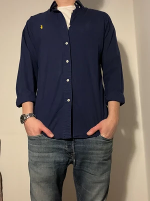 Mörkblå skjorta från Ralph Lauren - Snygg mörkblå skjorta från Ralph Lauren med klassisk gul logga broderad på bröstet. Skjortan har vita knappar och långärmad design med uppvikta ärmar. Tillverkad i mjuk bomull som känns skön mot huden. Perfekt för dig som gillar stilrena och tidlösa plagg. Size S
