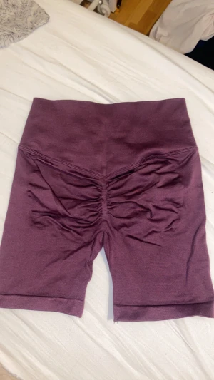 Mörklila scrunch shorts - Säljer ett par mörklila scrunch shorts med tight passform. De är tillverkade i ett mjukt och stretchigt syntetmaterial, perfekta för träning. Baksidan har snygg scrunch-detalj som framhäver formen. Storlek S men passar även XS