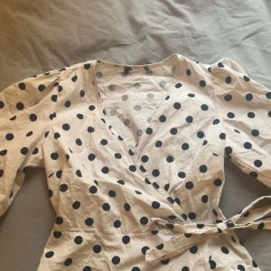 Prickig omlottblus med puffärm - Säljer en vit omlottblus med svarta prickar och puffiga trekvartsärmar. Blusen har v-ringning och knyt i midjan för en snygg siluett. Kommer inte ihåg märket tyvärr, storlek XS