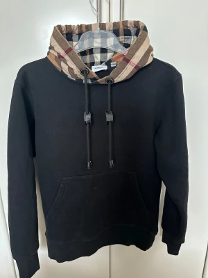 Burberry hoodie - Svart hoodie från Burberry med klassisk rutig huva i beige, svart och brunt. Tröjan har snörning vid huvan, känguruficka framtill och ribbade muddar. 