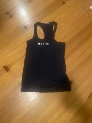 Svart linnetopp med MARGO-tryck - Svart linnetopp med racerback och silvrigt MARGO-tryck framtill. Perfekt för dig som gillar enkel stil med en touch av bling. Tunn och mjuk bomullskänsla, passar till både jeans och shorts. Snyggt snitt som visar axlarna.