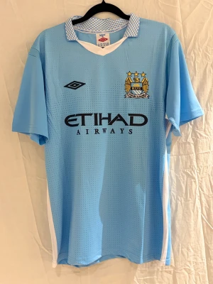 Manchester City Balotelli 45 tröja - Säljer en ljusblå Manchester City matchtröja med Balotelli och nummer 45 på ryggen. Tröjan har korta ärmar, vit krage med rutigt mönster och broderat klubbmärke på bröstet. Materialet är lätt och ventilerande polyester med små prickar.