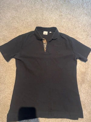 Svart Burberry skjorta med rutig detalj - Svart skjorta från Burberry i storlek L med klassisk krage och korta ärmar sitter bra på m även . Snygg rutig knappslå i beige, svart och rött som ger en ikonisk touch. Diskret broderad logga på bröstet. Skön bomullskvalitet som känns chill och stilren.