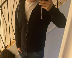 Marinblå hoodie från Polo Ralph Lauren - Snygg marinblå hoodie från Polo Ralph Lauren med gul broderad logga på bröstet. Tröjan har vit snörning i huvan, dragkedja framtill och två fickor. Tillverkad i mjuk bomull och har en klassisk, normal passform. Perfekt för chill dagar och streetwear. Är i storlek M men sitter mer som S