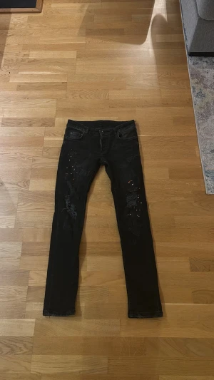 Dsquared2 jeans svarta - Svarta jeans från Dsquared2 med skinny/slim passform. Byxorna har slitningar och små färgstänk på framsidan för en edgy look. Klassisk femficksmodell med Dsquared2-logga vid gylfen och svart patch bak i midjan. Tillverkade i stretchig denim för extra komfort.