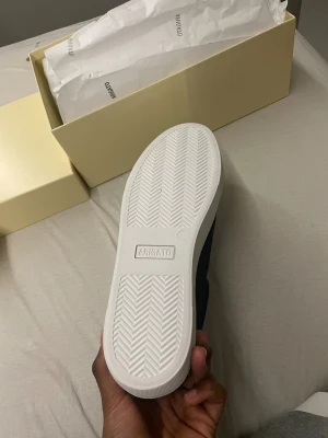 Svarta sneakers Axel Arigato mocka - Svarta sneakers från Axel Arigato i mocka med vit platt sula och elastiskt band över vristen. Minimalistisk design med rund tå och diskret logga på tungan och sulan. Perfekt för dig som gillar stilrena och cleana skor. Helt nya och oanvända.