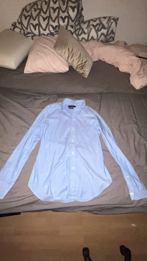 Blå slim fit skjorta från Ralph Lauren - Snygg ljusblå skjorta från Ralph Lauren i slim fit-modell. Klassisk design med knappar hela vägen, lång ärm och ikonisk broderad logga på bröstet. Skjortan är gjord i mjuk bomull och har en stilren krage som ger en clean look.