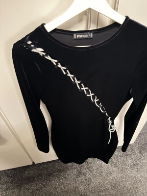 Svart sammetsklänning med snörning - Supersnygg svart sammetsklänning från FB Sister med lång ärm och tight passform. Klänningen har en cool snörningsdetalj i silver som går diagonalt över framsidan och ger en unik look. Perfekt för dig som vill sticka ut med stil. L men skulle säga passa mer till m eller s