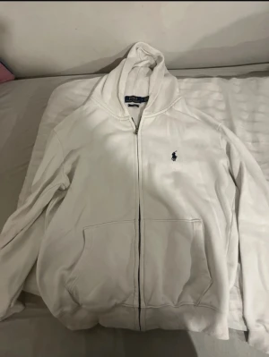 Vit hoodie från Polo Ralph Lauren - Vit hoodie från Polo Ralph Lauren i storlek L, med klassisk blå logga på bröstet. Tröjan har huva, dragkedja och två stora fickor framtill. Materialet är mjuk bomull och passformen är normal. Perfekt för chill dagar och enkel att matcha med jeans. PASSAR M