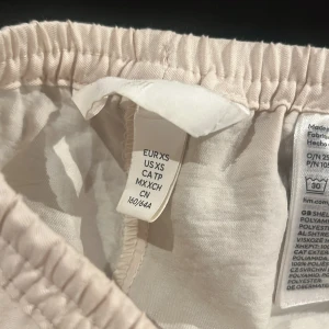Beiga linnebyxor med snörning från H&M - Säljer ett par beiga linnebyxor från H&M i storlek XS. Byxorna har en avslappnad wide passform, hög midja med resår och snörning framtill. Materialet är mjukt och luftigt, perfekt för chill dagar. Färgen är ljus beige och de har raka ben.