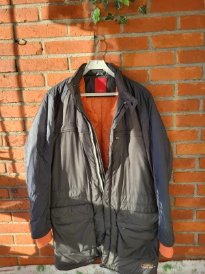 Grå jacka från J.Lindeberg - Säljer en grå pufferjacka från J.Lindeberg med orange quiltat foder. Jackan har flera praktiska fickor, hög krage med knapp och dragkedja framtill. Materialet är polyester och jackan har en loose passform, perfekt för kalla dagar.