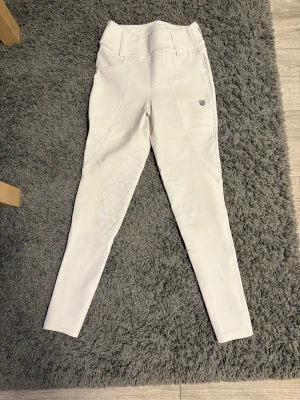 Vita ridbyxor med hög midja från Maya delorez  - Snygga vita ridbyxor med hög midja och smal passform. Byxorna har silikongrepp på insidan av benen för extra fäste i sadeln och praktiska bakfickor med lock. Perfekta för dig som vill ha stilrena och funktionella ridbyxor. Dem ser ut som nya och har inga tecken på användning.