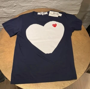 Blå Comme des Garçons Play t-shirt - Snygg mörkblå t-shirt från Comme des Garçons Play med stort vitt hjärta och rött hjärtdetalj med ögon på bröstet. Klassisk rund hals och korta ärmar. Tillverkad i 100% bomull, mjuk och skön kvalitet. Perfekt för dig som gillar ikoniska prints och streetstyle.