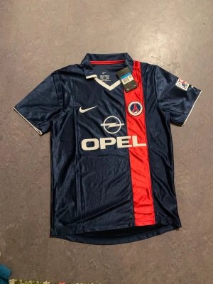 PSG Ronaldinho 21 Nike fotbollströja - Säljer en klassisk Paris Saint-Germain fotbollströja med Ronaldinho och nummer 21 på ryggen. Tröjan är marinblå med en röd vertikal rand, vita detaljer och PSG-logga på bröstet. Tillverkad i glansigt polyester, har krage och korta ärmar. Perfekt för dig som älskar retro och fotbollshistoria.