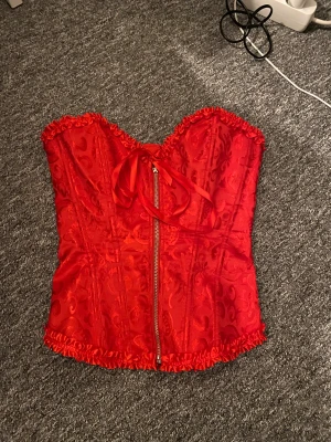 Röd korsett med snörning och dragkedja - Säljer en röd korsett med snyggt mönster och glansig finish. Toppen har snörning i ryggen, dragkedja framtill och volangkant upptill och nedtill. Materialet känns lyxigt och har en satinliknande look. Använd en gång.