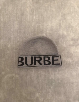Grå Burberry mössa med logga - Snygg grå mössa från Burberry med bred kant och stor svart logotyp runt om. Mjuk och stickad i ull, perfekt för att hålla värmen och samtidigt se stilren ut. Klassisk form som passar till de flesta outfits och har en exklusiv känsla.
