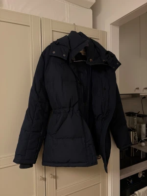 Mörkblå dunjacka från Boomerang - Snygg mörkblå dunjacka från Boomerang med huva, knappar och dragkedja. Jackan har quiltad design, stora fickor och justerbar midja. Perfekt för kalla dagar med sin fluffiga och värmande känsla.