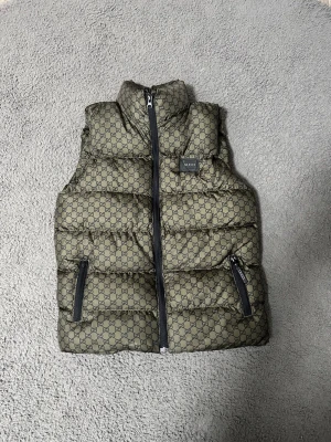 Zara pufferväst med GG-mönster - Säljer en fet Zara pufferväst i olivgrön färg med all-over GG-logga. Västen har hög krage, två dragkedjefickor och svart innerfoder. Materialet är syntet och polyester, perfekt för att maxa din stil när det är kallt ute. Dragkedja framtill och tydlig Zara patch på bröstet.