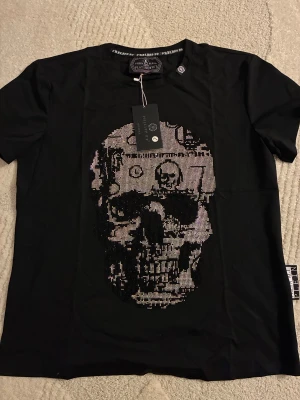 Svart t-shirt med döskalle från Philipp Plein - Svart t-shirt från Philipp Plein med stor glittrig döskalle framtill. T-shirten har korta ärmar och är gjord i mjuk bomull. Märkesdetaljer syns i nacken och på sidan. Perfekt för dig som gillar edgy och exklusiv streetstyle.