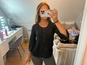 Mörkgrå ribbad blus med knappar - Säljer en mörkgrå ribbad blus med rund hals och tre stora dekorativa knappar framtill. Blusen har trekvartsärm och lätt utsvängd passform som ger en snygg siluett. Perfekt för dig som gillar stilrena detaljer och mjuka material.