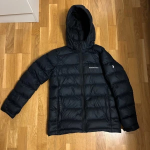 Marinblå Dunjacka från Peak Performance - Säljer en marinblå dunjacka från Peak Performance i mycket bra skick. Jackan är perfekt för kalla dagar med sin pufferstil och långa ärmar. Den har en praktisk dragkedja och en huva för extra värme. Perfekt för både höst och vinter! Storlek 170, motsvar ungefär XS/S