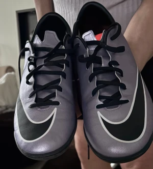 Nike fotbollsskor i silver och svart - Säljer ett par stilrena Nike fotbollsskor i silver med svarta detaljer och stora swoosh-loggor. Skorna har snörning, platt sula och rund tå, perfekt för inomhusspel. Materialet är syntet och designen är riktigt clean med en sportig vibe.