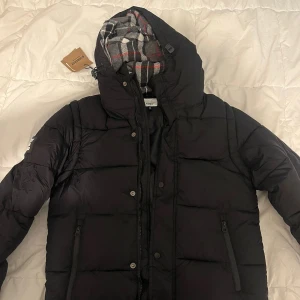 Svart pufferjacka från Burberry finns i storlek M och S - Säljer en svart pufferjacka från Burberry med avtagbara ärmar så den kan användas som väst. Jackan har huva med rutigt foder i grått, vitt och rött, samt dragkedja och knappar framtill. Två fickor med dragkedja och stor Burberry-logga på ärmen.