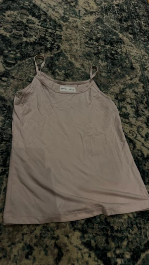 Rosa linnetopp från Lefties - Säljer en enkel och stilren rosa linnetopp från Lefties i storlek M. Toppen har smala axelband och ärmlös design, perfekt för varma dagar. Materialet är mjukt och lätt polyester som känns skönt mot huden. Passformen är normal och toppen är enfärgad utan mönster.