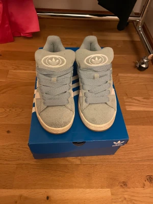Adidas Campus ljusblå sneakers mocka - Säljer ett par Adidas Campus sneakers i ljusblå mocka med klassiska vita ränder på sidan och Adidas-logga på plösen och hälen. Skorna har rund tå, platt sula och breda snören för en cool vibe. Perfekt för dig som gillar retrostil och streetwear. Säljer dem för minst 275 kr❤️