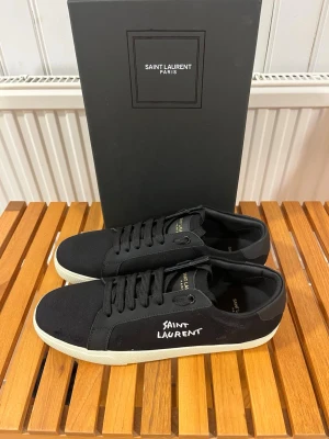  Saint Laurent skor - Svarta skor från Saint Laurent med vit sula och handskriven logga på sidan. Skorna har snörning, rund tå och är tillverkade i textil och skinn. Snygg minimalistisk design med gulddetaljer på plösen. Perfekta för dig som gillar stilrena och exklusiva skor. Skorna är i bra skick och är storlek 44