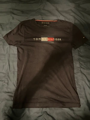 Svart t-shirt Tommy Hilfiger S - Svart t-shirt från Tommy Hilfiger i storlek S, tillverkad i ekologisk bomull. Framsidan har ett stilrent tryck med logotypen i vitt, rött och blått. Klassisk rund hals och korta ärmar. Perfekt för dig som gillar snygga och enkla märkesplagg.
