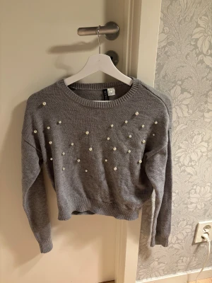 Grå stickad tröja med pärlor Divided - Snygg grå stickad tröja från Divided i storlek XS. Tröjan har långa ärmar och är dekorerad med pärlor framtill för en cool och trendig look. Ribbad kant vid hals, ärmslut och nederkant. Perfekt för dig som gillar detaljer!