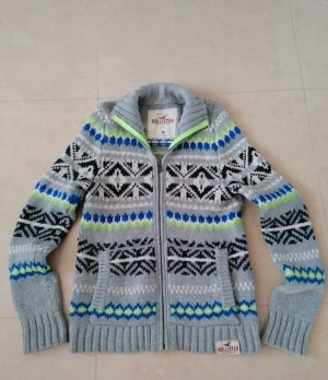 Grå mönstrad kofta från Hollister - Säljer en grå stickad kofta från Hollister med dragkedja och snyggt mönster i svart, blått och limegrönt. Koftan har ribbade muddar, hög krage och två fickor framtill. Materialet är en mix av bomull, nylon och ull. Perfekt för kyliga dagar!